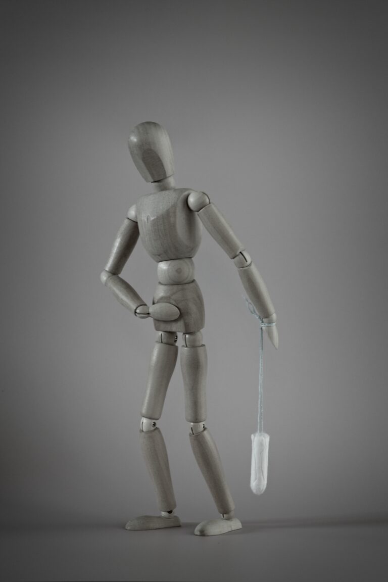 A wooden figure looks downward uncomfortably, holding a tampon behind its back, symbolizing the last menstruation and the transition into the unknown.