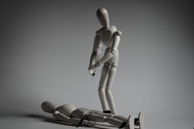 A wooden figure lies restrained as another approaches with a syringe, reflecting the themes of animal testing and power imbalance in Beproevingen – Lara Part 1.