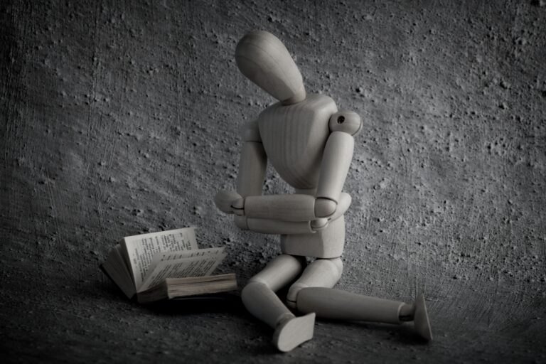 Een houten figuur zit ineengedoken tegen een kale muur. Naast hem ligt een open boek. De sfeer is teruggetrokken en beklemmend.